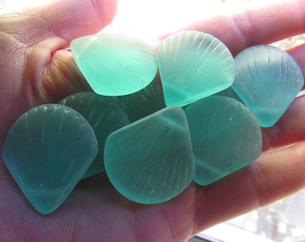 May include: A collection of six teal blue glass sea shells with a fan-shaped design. The shells are smooth and translucent.
