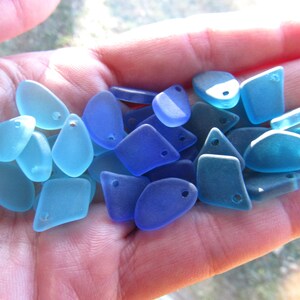 May include: A collection of small, smooth, translucent glass pieces in various shades of blue, ranging from light aqua to deep cobalt. Each piece features a single drilled hole, making them suitable for use as beads or jewelry components.