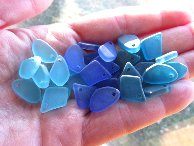 May include: A collection of small, smooth, translucent glass pieces in various shades of blue, ranging from light aqua to deep cobalt. Each piece features a single drilled hole, making them suitable for use as beads or jewelry components.
