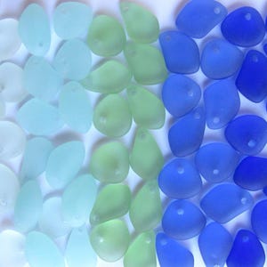 May include: An assortment of sea glass pieces in a gradient of colors, from white to light blue, green, and deep blue. Each piece has a smooth, frosted appearance and a small hole for stringing.