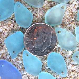 May include: A collection of frosted, light blue and dark blue sea glass pieces, each featuring a single pre-drilled hole for jewelry making. A silver-colored United States dime is positioned in the center for scale, measuring 0.705 inches in diameter.