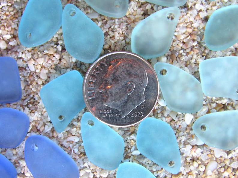 May include: A collection of frosted, light blue and dark blue sea glass pieces, each featuring a single pre-drilled hole for jewelry making. A silver-colored United States dime is positioned in the center for scale, measuring 0.705 inches in diameter.