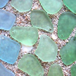 May include: Close-up of sea glass pieces in shades of blue and green, with small holes for jewelry making. The sea glass is scattered on a bed of sand and small pebbles, creating a natural, beachy aesthetic.