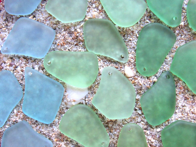 May include: Close-up of sea glass pieces in shades of blue and green, with small holes for jewelry making. The sea glass is scattered on a bed of sand and small pebbles, creating a natural, beachy aesthetic.