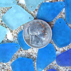 May include: A collection of sea glass pieces in various shades of blue and turquoise, scattered on sand. A U.S. quarter coin is in the center, providing a size reference. The sea glass pieces have a smooth, frosted appearance and are of different shapes.