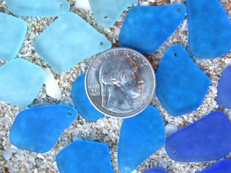 May include: A collection of sea glass pieces in various shades of blue and turquoise, scattered on sand. A U.S. quarter coin is in the center, providing a size reference. The sea glass pieces have a smooth, frosted appearance and are of different shapes.