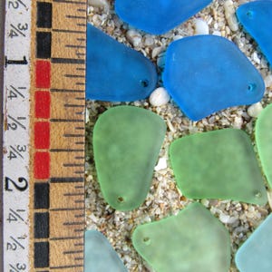 May include: A collection of sea glass pieces in shades of blue and green, with small holes, resting on a sandy surface. A wooden ruler with inch measurements is visible along the left side of the image.