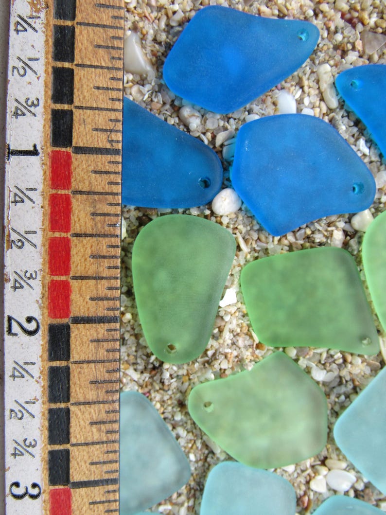 May include: A collection of sea glass pieces in shades of blue and green, with small holes, resting on a sandy surface. A wooden ruler with inch measurements is visible along the left side of the image.