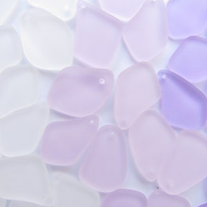May include: A collection of frosted glass pendants in shades of white, lavender, and purple. Each pendant has a smooth, irregular shape and a small hole for stringing. The pendants are scattered across a white surface.