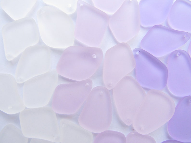 May include: A collection of frosted glass pendants in shades of white, lavender, and purple. Each pendant has a smooth, irregular shape and a small hole for stringing. The pendants are scattered across a white surface.