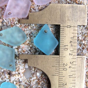 May include: A collection of frosted, translucent sea glass pendants in shades of light blue and pale pink, each featuring a single drilled hole. A brass caliper is positioned over one blue diamond-shaped pendant, showing a width of 0.75 inches.