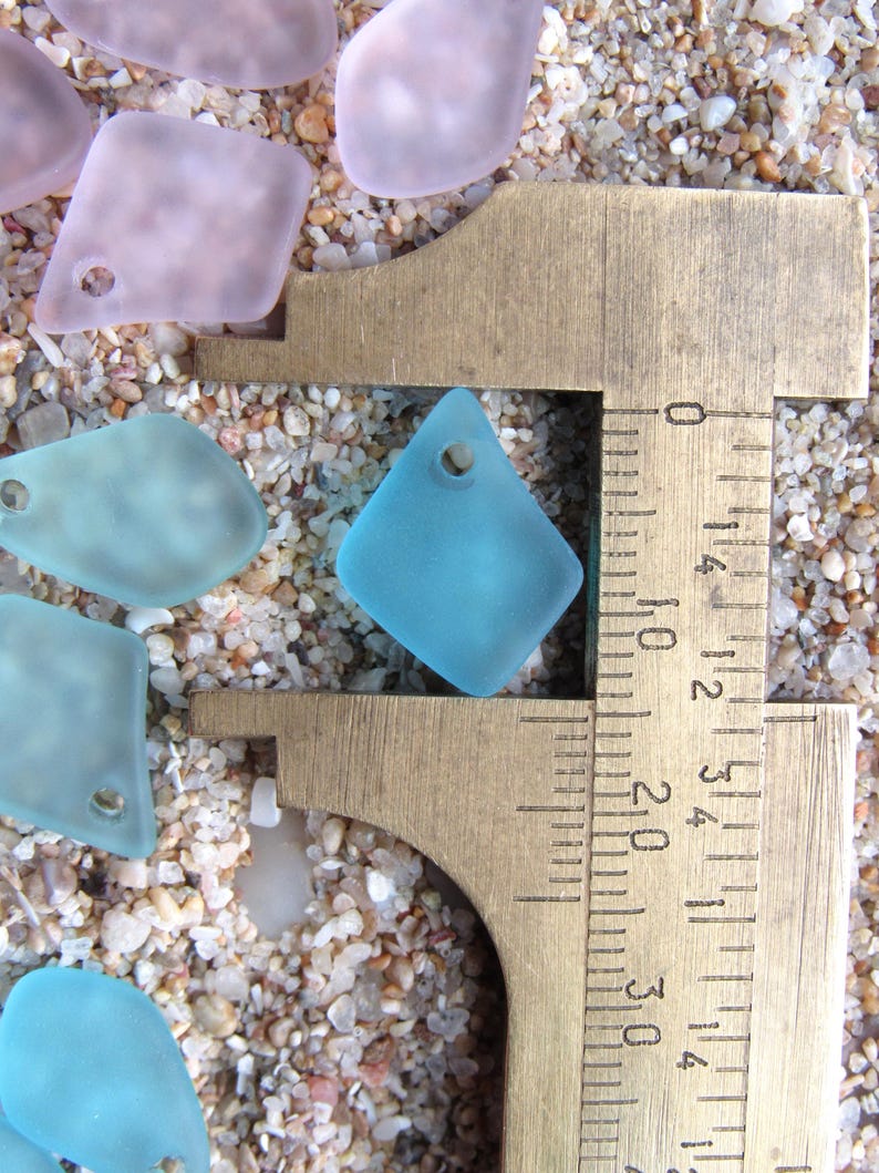 May include: A collection of frosted, translucent sea glass pendants in shades of light blue and pale pink, each featuring a single drilled hole. A brass caliper is positioned over one blue diamond-shaped pendant, showing a width of 0.75 inches.