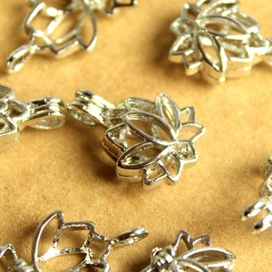 May include: Close-up of several silver-colored metal lotus flower-shaped jewelry findings. Each finding has a loop for attaching to a chain or other jewelry component. The findings are scattered on a light brown surface.