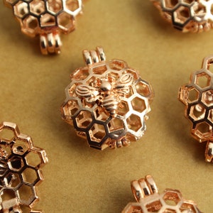Può includere: Ciondoli a forma di favo d'oro rosa con un motivo a forma di ape. I ciondoli presentano un design dettagliato a celle esagonali e una piccola ape al centro. Hanno un anello nella parte superiore per attaccare una catena.