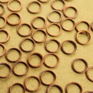 May include: A close-up of a group of copper jump rings, scattered on a brown surface. The rings are all the same size and shape, and they are shiny and metallic.