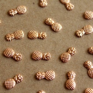 May include: A collection of gold-toned pineapple shaped charms. The charms are scattered on a brown surface.