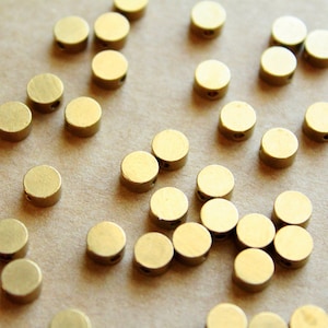 May include: A close-up of a group of gold-colored metal beads. The beads are round and have a smooth, shiny finish. They are scattered on a light brown surface.