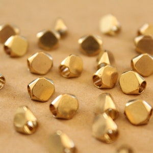 May include: A collection of small, geometric beads with a metallic gold finish. Each bead has multiple flat sides and a hole running through the center, suitable for stringing. The beads are scattered on a neutral-toned surface.