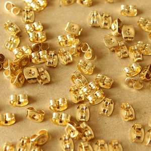May include: A collection of small, shiny gold-colored earring backs scattered on a light brown surface. The earring backs are various shapes and sizes, with a smooth, reflective finish. The image is well-lit, highlighting the metallic sheen of the jewelry findings.