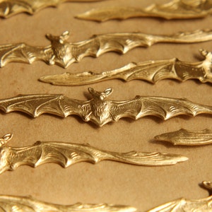 May include: Close-up of several gold-colored bat-shaped embellishments. The bats have detailed wings and bodies, and are arranged on a light brown surface. The bats are likely for crafting or decorative purposes.