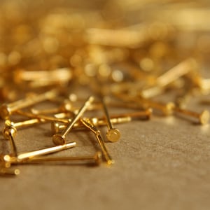 May include: A close-up of a pile of gold-colored metal pins. The pins are scattered on a brown surface.