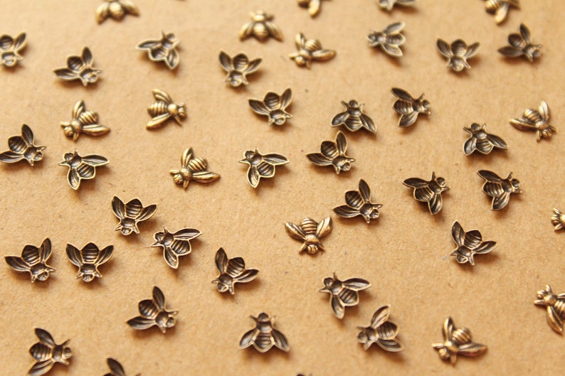 May include: A collection of small, gold-toned metal bee charms scattered on a brown surface. The bees are all facing different directions and have a detailed design.