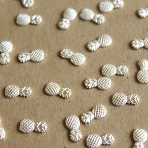 May include: A collection of silver pineapple charms, scattered on a brown surface. The charms are small and detailed, with a textured pineapple design.