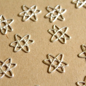 May include: A collection of silver-colored metal charms shaped like atoms. The charms are scattered on a brown surface.