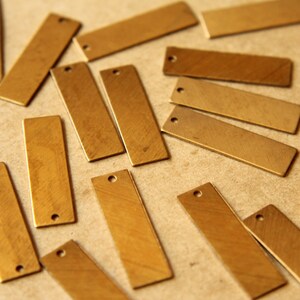 May include: A collection of rectangular brass tags with a small hole at one end. The tags are scattered across a neutral surface, showcasing their warm, metallic color and simple design. Ideal for jewelry making or crafting.
