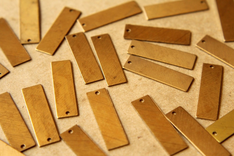 May include: A collection of rectangular brass tags with a small hole at one end. The tags are scattered across a neutral surface, showcasing their warm, metallic color and simple design. Ideal for jewelry making or crafting.