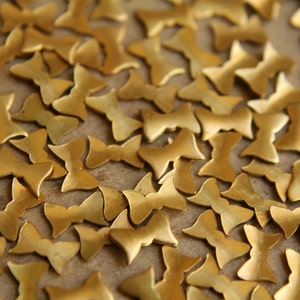 May include: A close-up image of a large quantity of gold-colored metal butterfly shapes. The shapes are scattered on a brown surface.