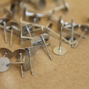 May include: A close-up of a pile of silver earring posts with flat backs. The posts are scattered on a brown surface.
