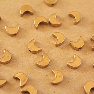 May include: A collection of gold crescent moon shaped beads. The beads are scattered on a brown surface.