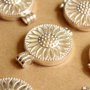 Könnte beinhalten: Sechs silberne Anhänger mit Sonnenblumen-Design. Die Anhänger haben eine Scharnieröffnung und eine kleine Schlaufe zum Anbringen einer Kette.