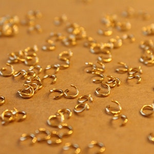 May include: A close-up of a large quantity of small, gold-colored jump rings. The rings are scattered on a brown surface.