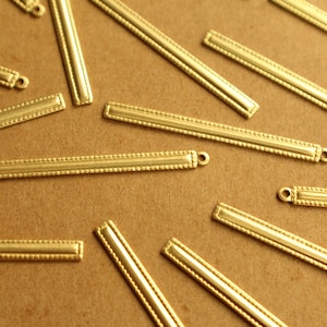 May include: Close-up of several gold-colored metal bar components with a rectangular shape. Each bar features a beaded border and a small loop at one end. The components are scattered on a brown surface.