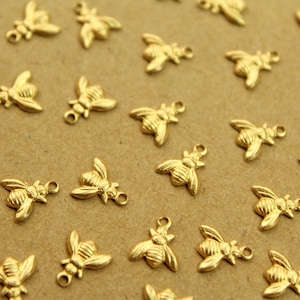 May include: A collection of gold-toned bee charms, scattered on a brown surface. Each charm features a detailed bee design with raised wings and a textured body.