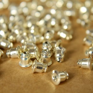 May include: A close-up of a pile of silver earring backs. The backs are small and round, with a flat surface on one side and a pointed tip on the other.