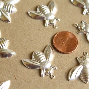 Puede incluir: Primer plano de un grupo de abalorios de abeja plateados, algunos con anillas de salto. Los abalorios est&aacute;n dispuestos sobre una superficie marr&oacute;n junto a una moneda de un c&eacute;ntimo de euro.