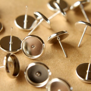 May include: A close-up of silver earring settings, also known as earring blanks, with a round, flat base and a post for attaching earrings. The settings are scattered on a brown surface.
