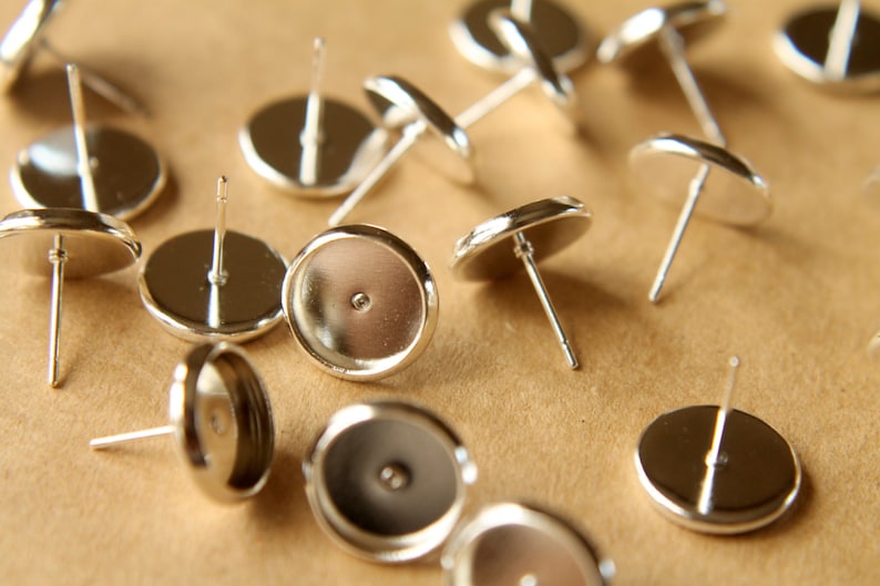 May include: A close-up of silver earring settings, also known as earring blanks, with a round, flat base and a post for attaching earrings. The settings are scattered on a brown surface.