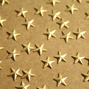 Puede incluir: Primer plano de numerosos pequeños adornos dorados en forma de estrella dispersos sobre una superficie marrón claro. Las estrellas están espaciadas uniformemente y tienen un brillo metálico, creando un patrón decorativo. Las estrellas miden aproximadamente 1,3 cm de diámetro.