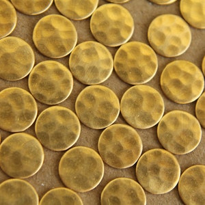 May include: Close-up of numerous small, hammered brass discs. Each disc is round with a textured surface and a golden hue. The discs are scattered across the frame, creating a pattern.
