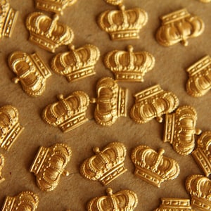May include: A close-up of a large quantity of gold-colored crown-shaped embellishments. The crowns are scattered on a brown surface.
