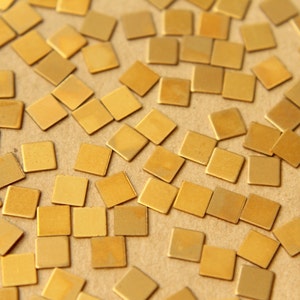 May include: A close-up image of a large quantity of small, square, gold-colored metal pieces scattered on a brown surface.