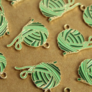 Puede incluir: Un grupo de diez abalorios de metal de color dorado con forma de ovillos de lana. La lana es verde y blanca con un lazo de metal dorado en la parte superior de cada abalorio.