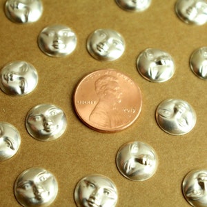 May include: A collection of silver-toned metal charms featuring a face on each side. The charms are arranged on a brown surface with a US penny for scale.