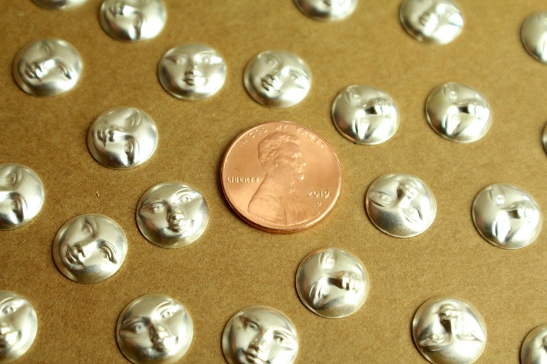 May include: A collection of silver-toned metal charms featuring a face on each side. The charms are arranged on a brown surface with a US penny for scale.