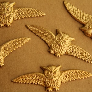 May include: Five gold-colored owl figurines with detailed wings and faces. The owls are arranged in a scattered pattern on a brown surface.