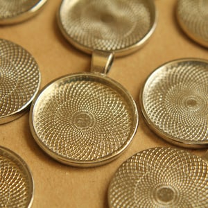May include: A close-up of several silver-toned round metal pendant trays with a textured swirl design. The trays are perfect for creating custom jewelry or crafts.